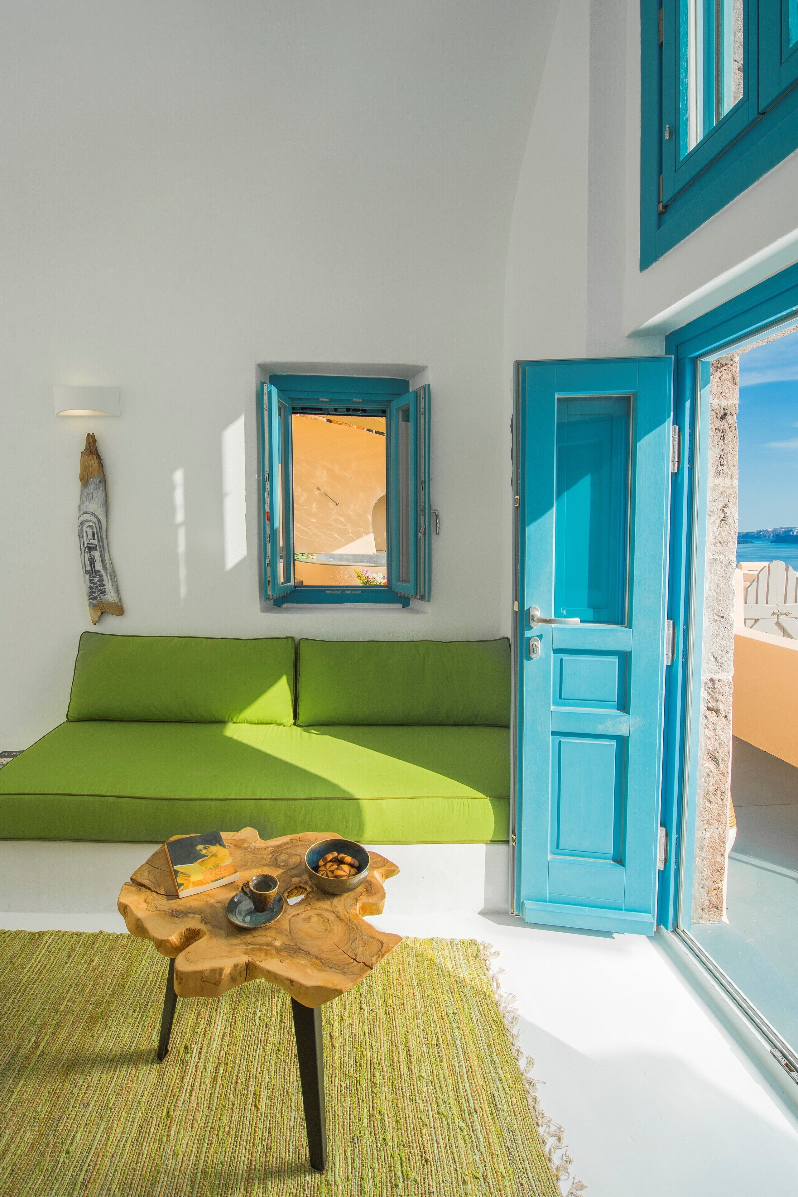 A bright room with a green sofa, a wooden coffee table with a book and a bowl, a small window, and a blue door leading to a balcony.