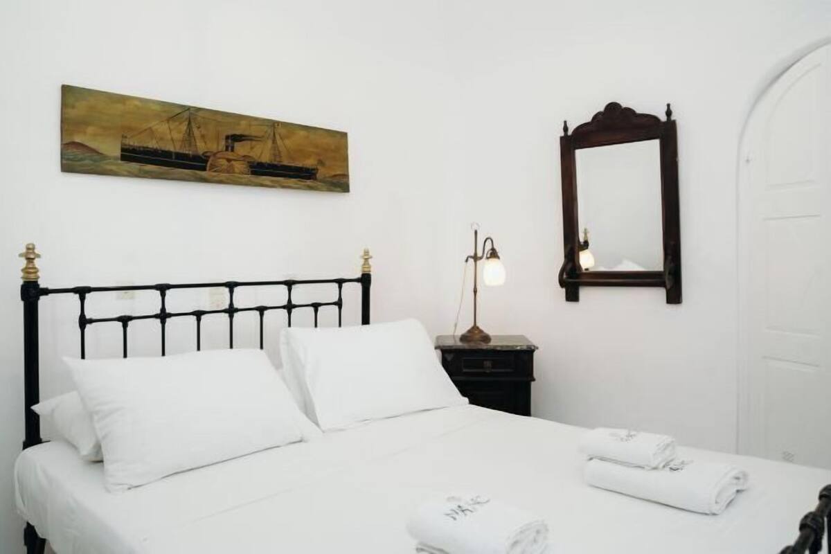 Una cama bien hecha con sábanas blancas, una lámpara de noche, un cuadro de un barco en la pared y un espejo.