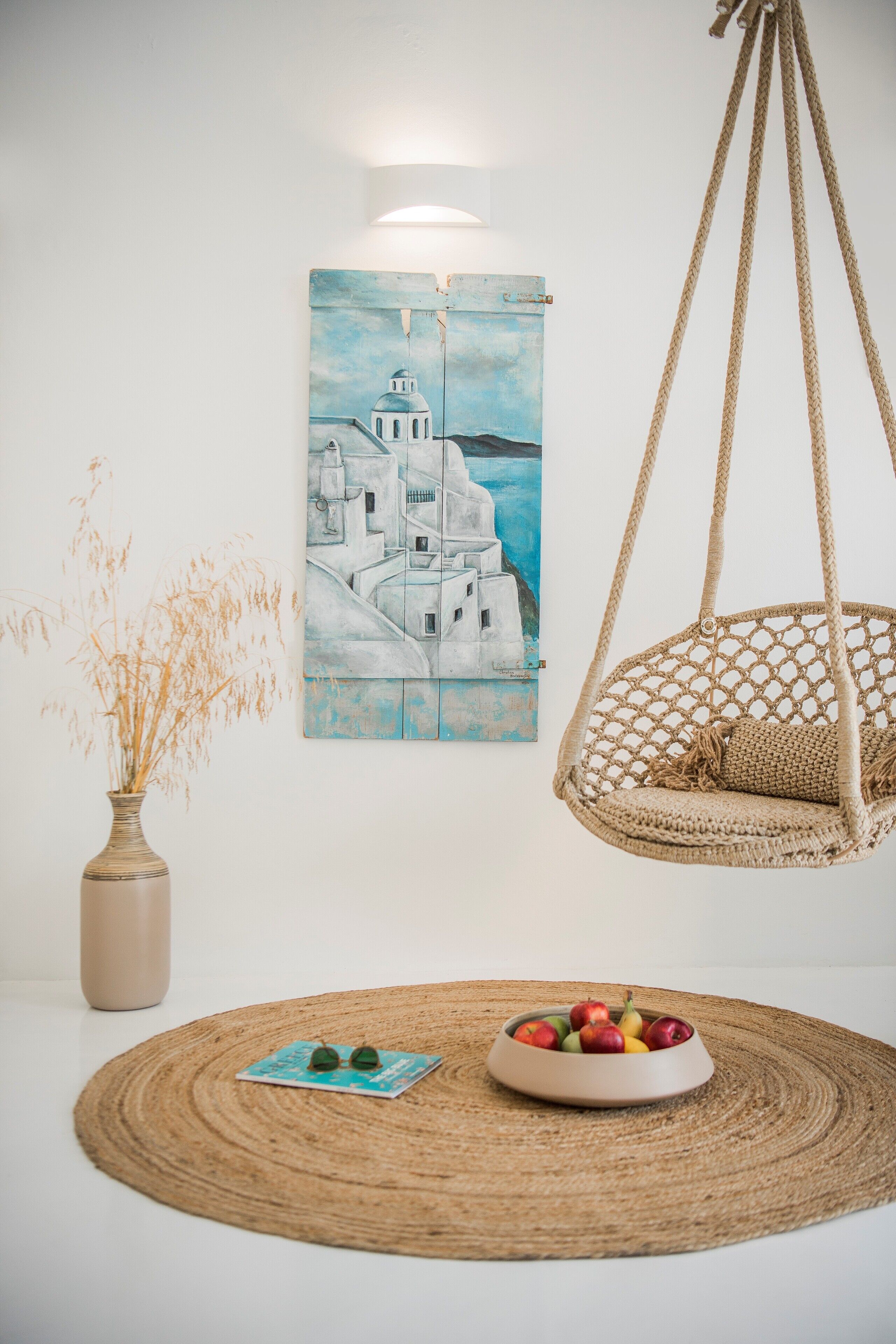 A room with a hanging chair, a vase with dried plants, and a painting of a coastal scene.