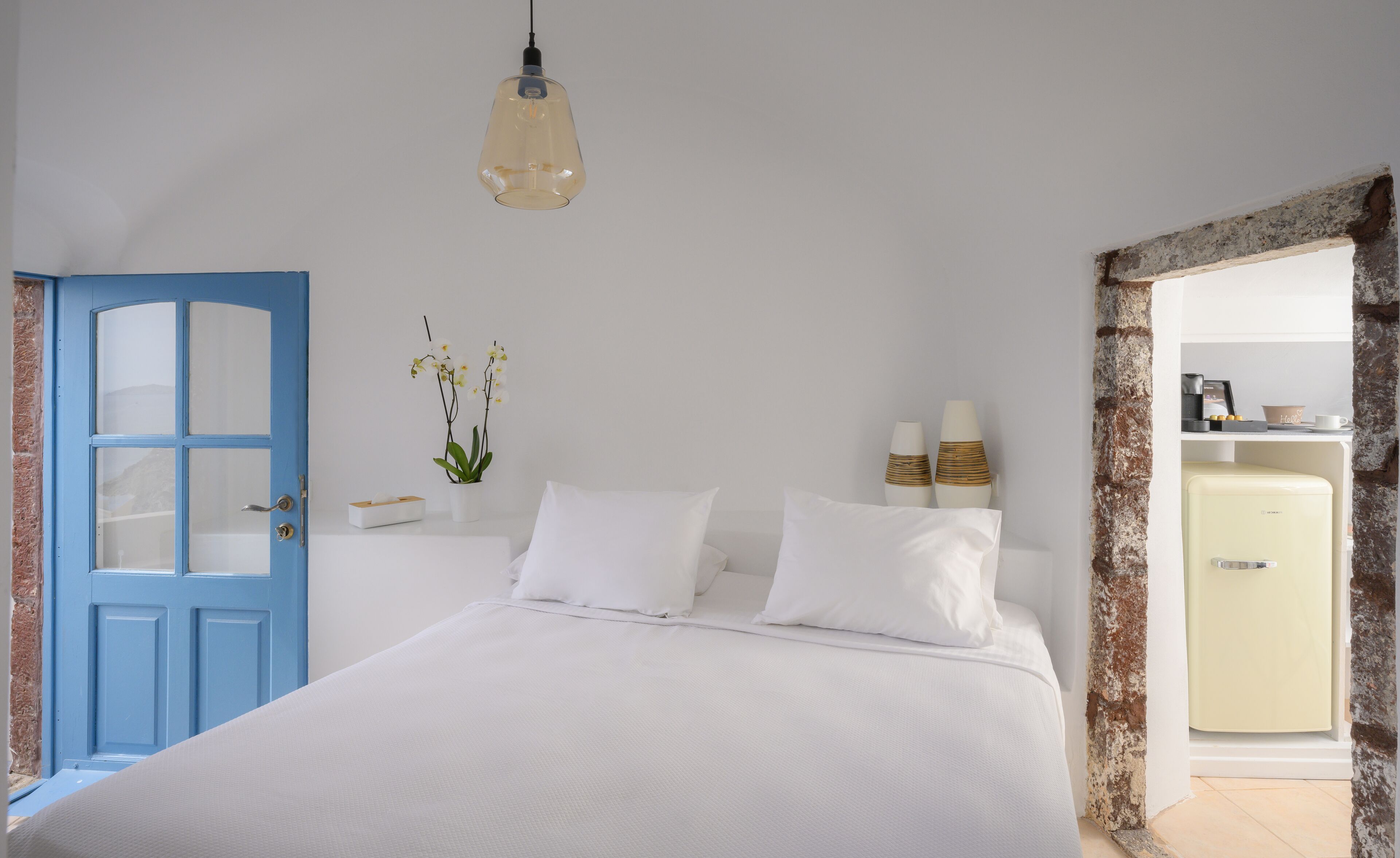 A bedroom with a bed, a blue door, a potted orchid, bedside lamps, and a vintage refrigerator.