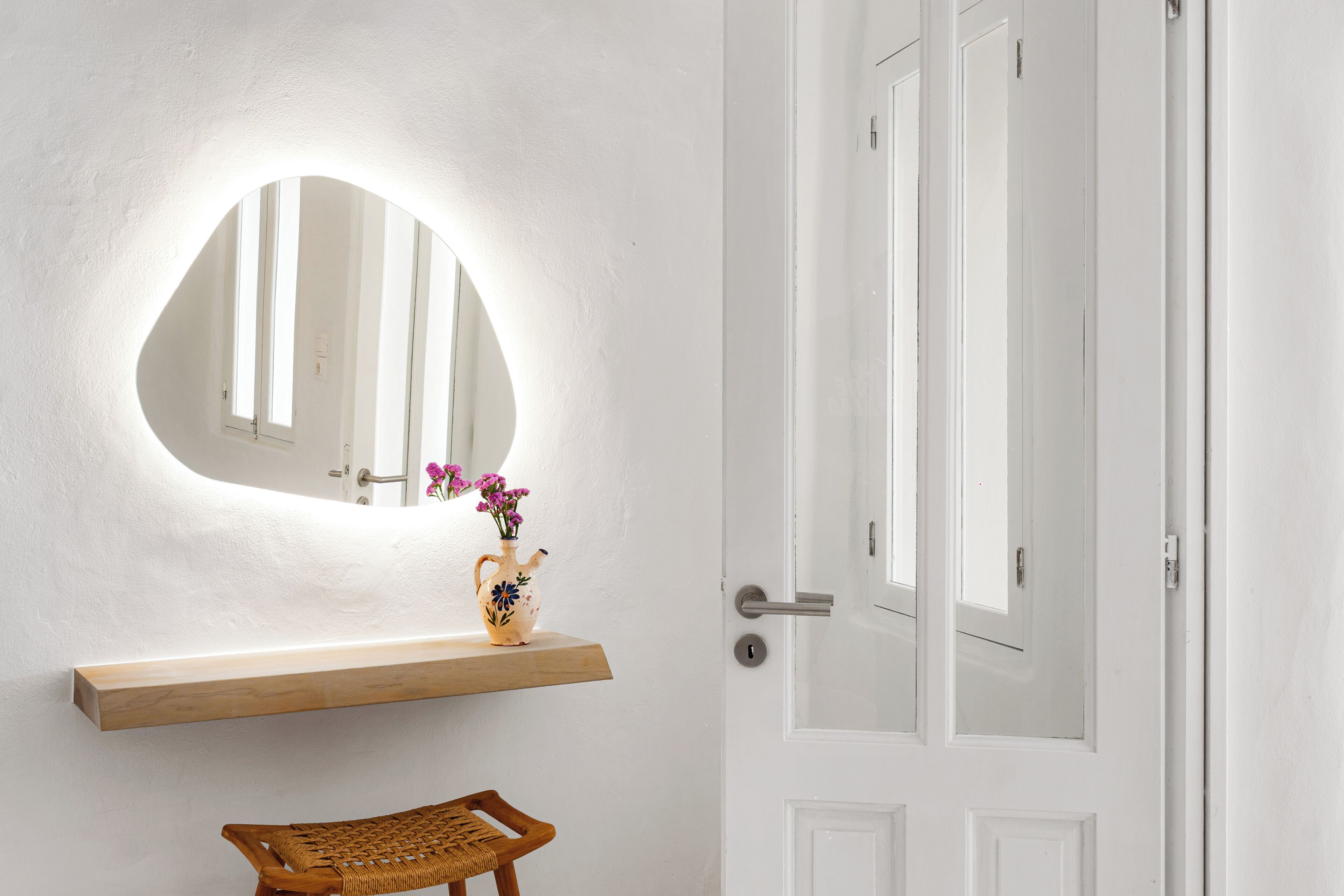 A bathroom with a white wall, a wooden shelf, a mirror, a door, and a wicker chair.