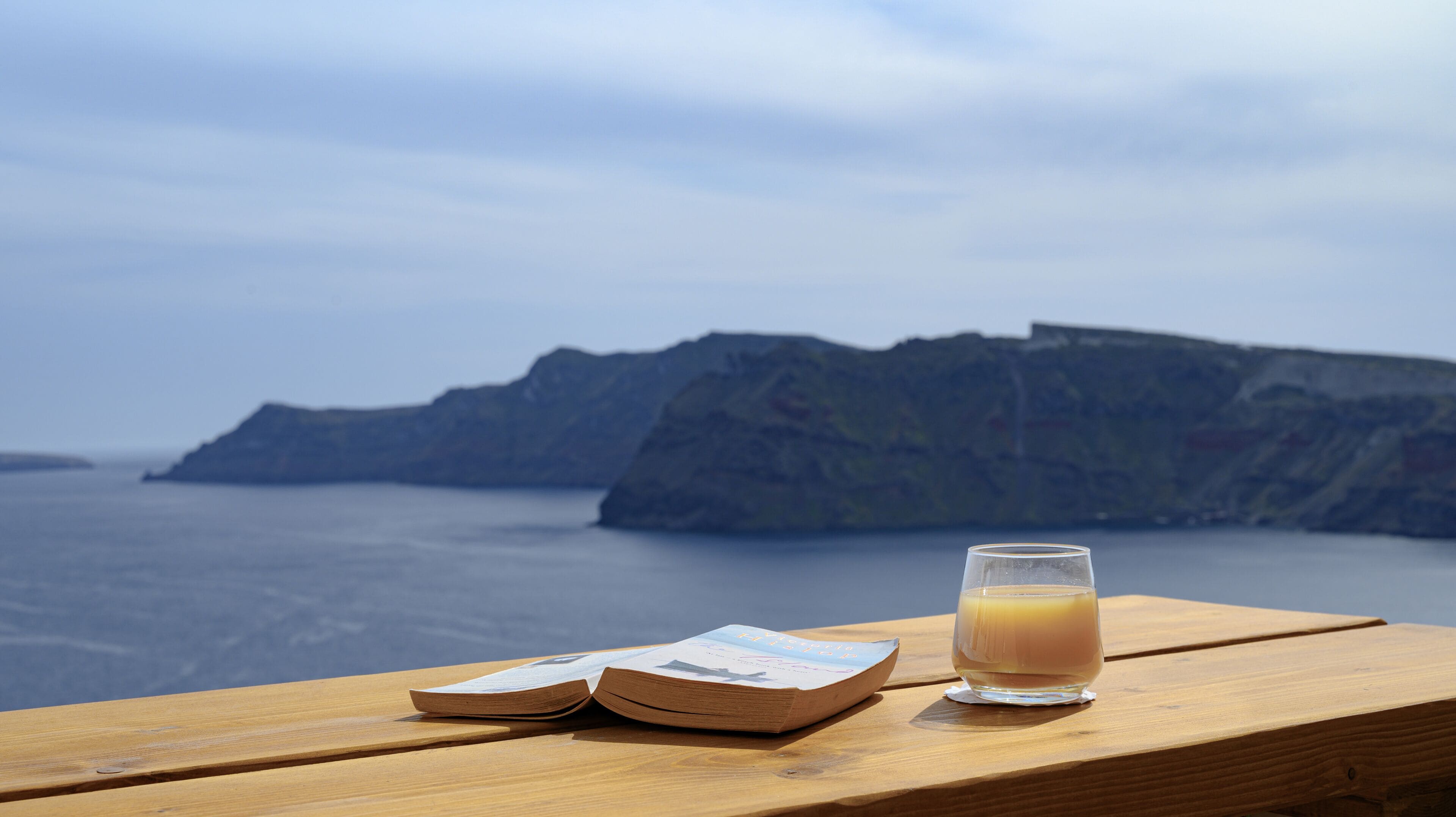Een houten tafel met een boek en een glas sinaasappelsap, met uitzicht op een wateroppervlak en een rotsachtige klif.