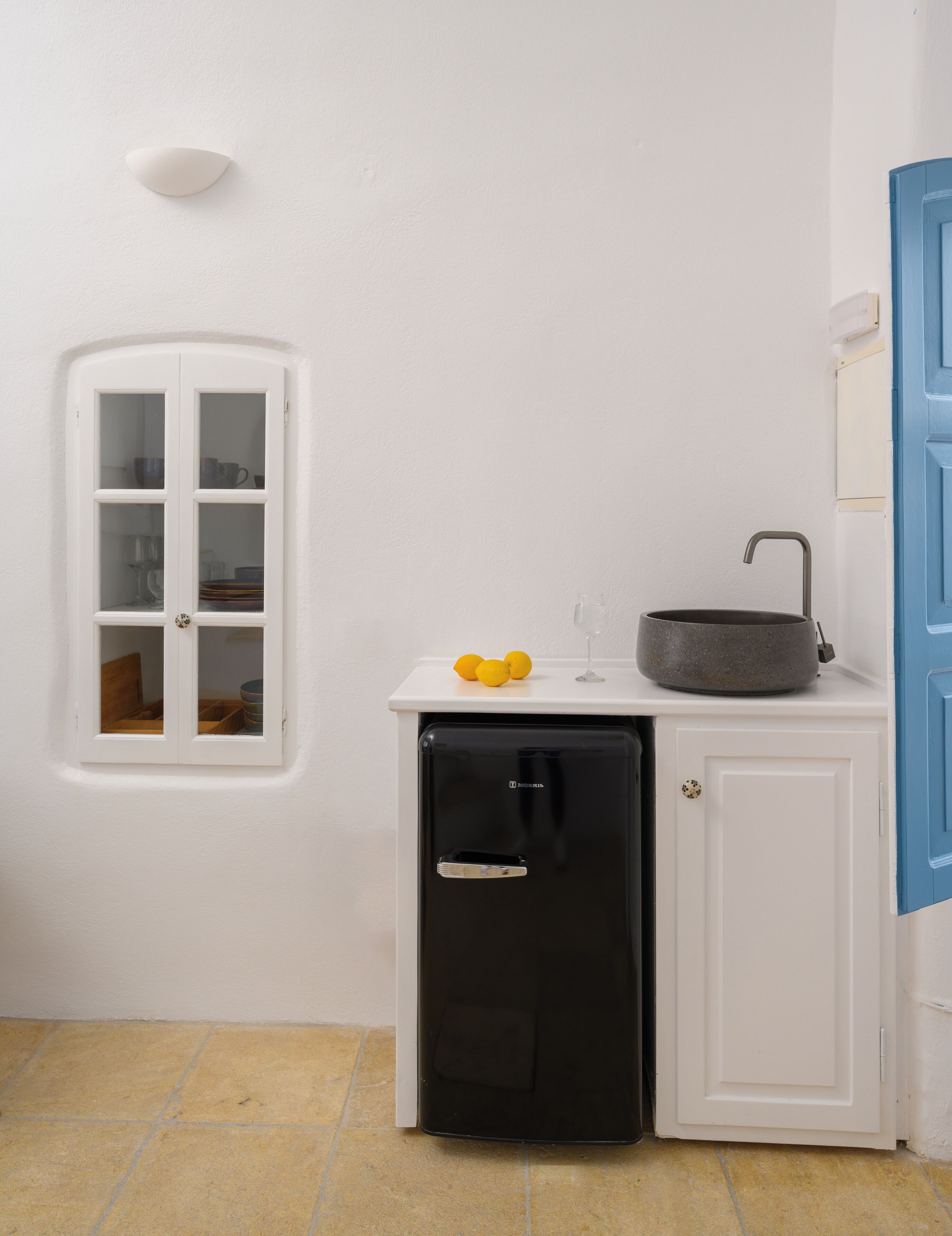 A compact kitchenette with a black refrigerator, a sink with a stone basin, and a small window with a view of a room with shelves.