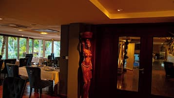 A dining area with tables and chairs, a wooden statue, and a view of greenery through large windows.