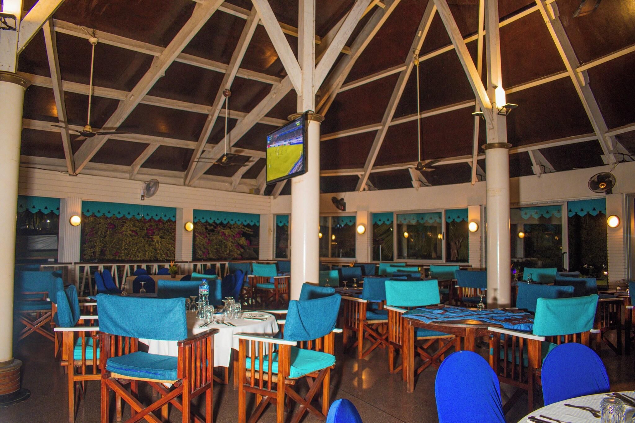 Un restaurante con mesas de madera y sillas azules, techo alto con vigas y una pantalla de televisión.