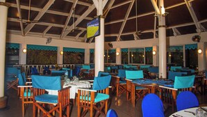 Un restaurant avec des tables en bois et des chaises bleues, un plafond haut avec des poutres apparentes et un écran de télévision.