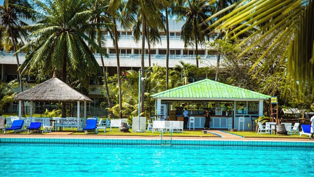 Une piscine bordée de chaises longues bleues, un abri à toit de chaume et un bâtiment surmonté d’un toit végétalisé.