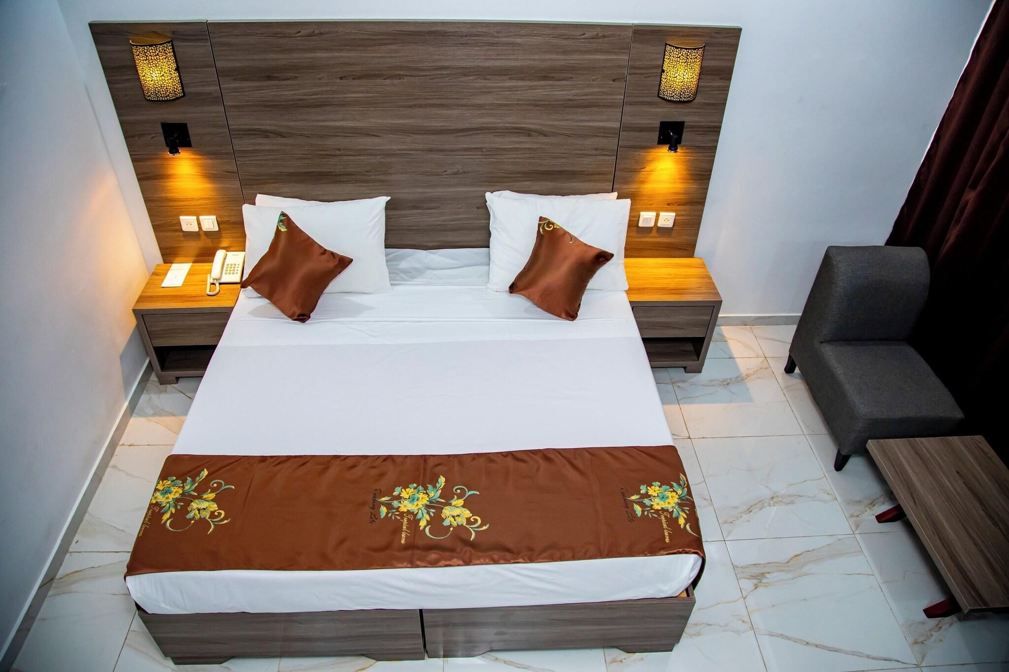 Una cama bien hecha con una colcha marrón, dos mesitas de noche con lámparas y un sillón gris.