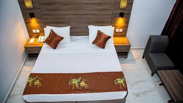 Una cama bien hecha con una colcha marrón, dos mesitas de noche con lámparas y un sillón gris.