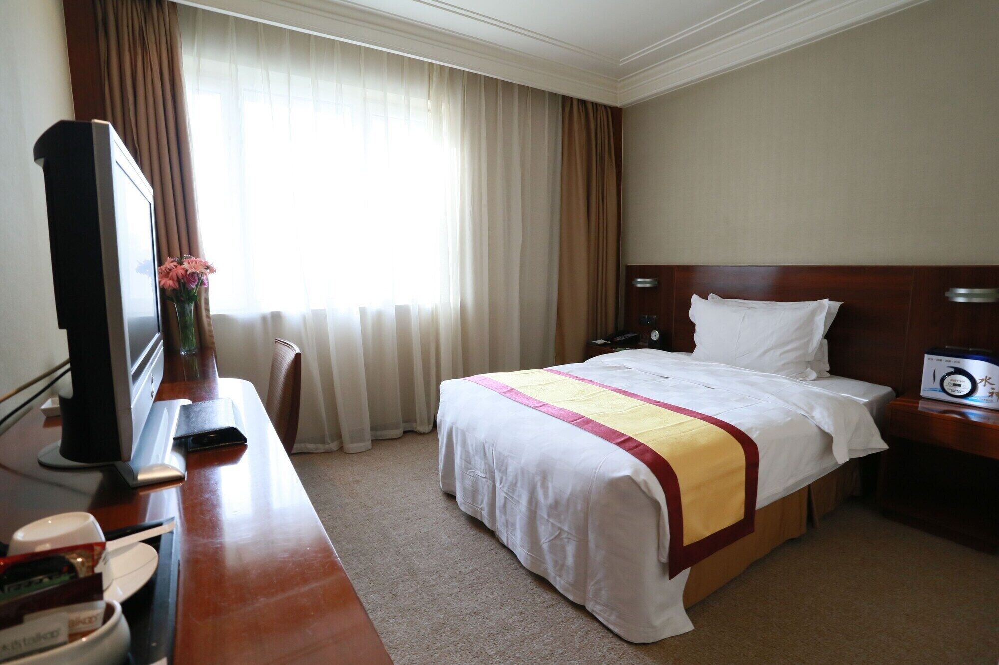A hotel room with a bed, a desk with a television, a bedside table with a clock, and a window with curtains.