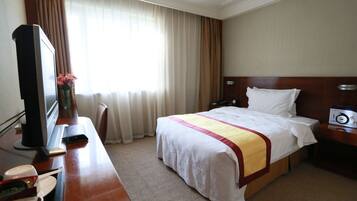 A hotel room with a bed, a desk with a television, a bedside table with a clock, and a window with curtains.