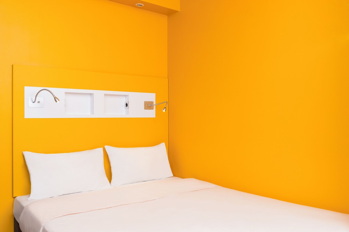 A bed with white bedding, a white headboard, and a white wall-mounted shelf with a towel and a light fixture.