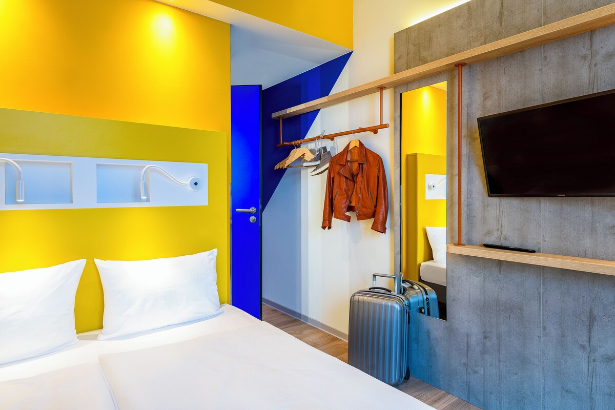 A hotel room with a bed, a television, a coat rack with a jacket, and a suitcase.