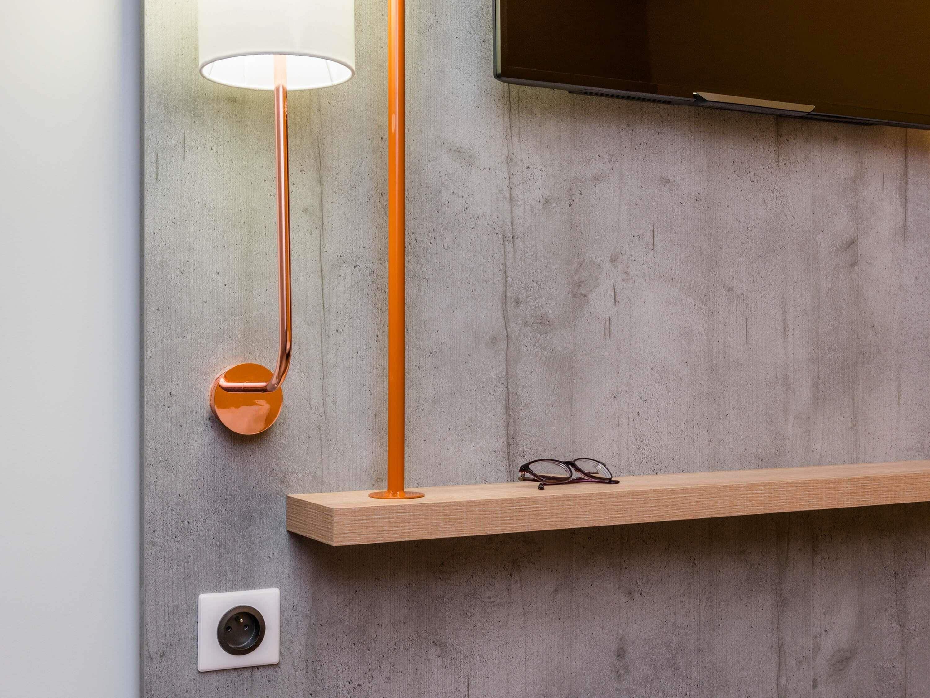Une pièce moderne avec un mur en béton, un téléviseur fixé au mur, une étagère en bois, une lampe sur pied et une prise électrique murale.