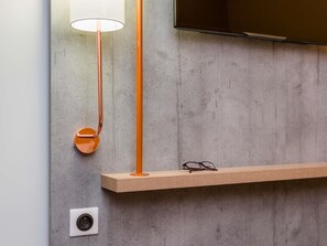 Una habitación moderna con pared de concreto, televisor montado, estante de madera, lámpara de pie y toma de corriente.