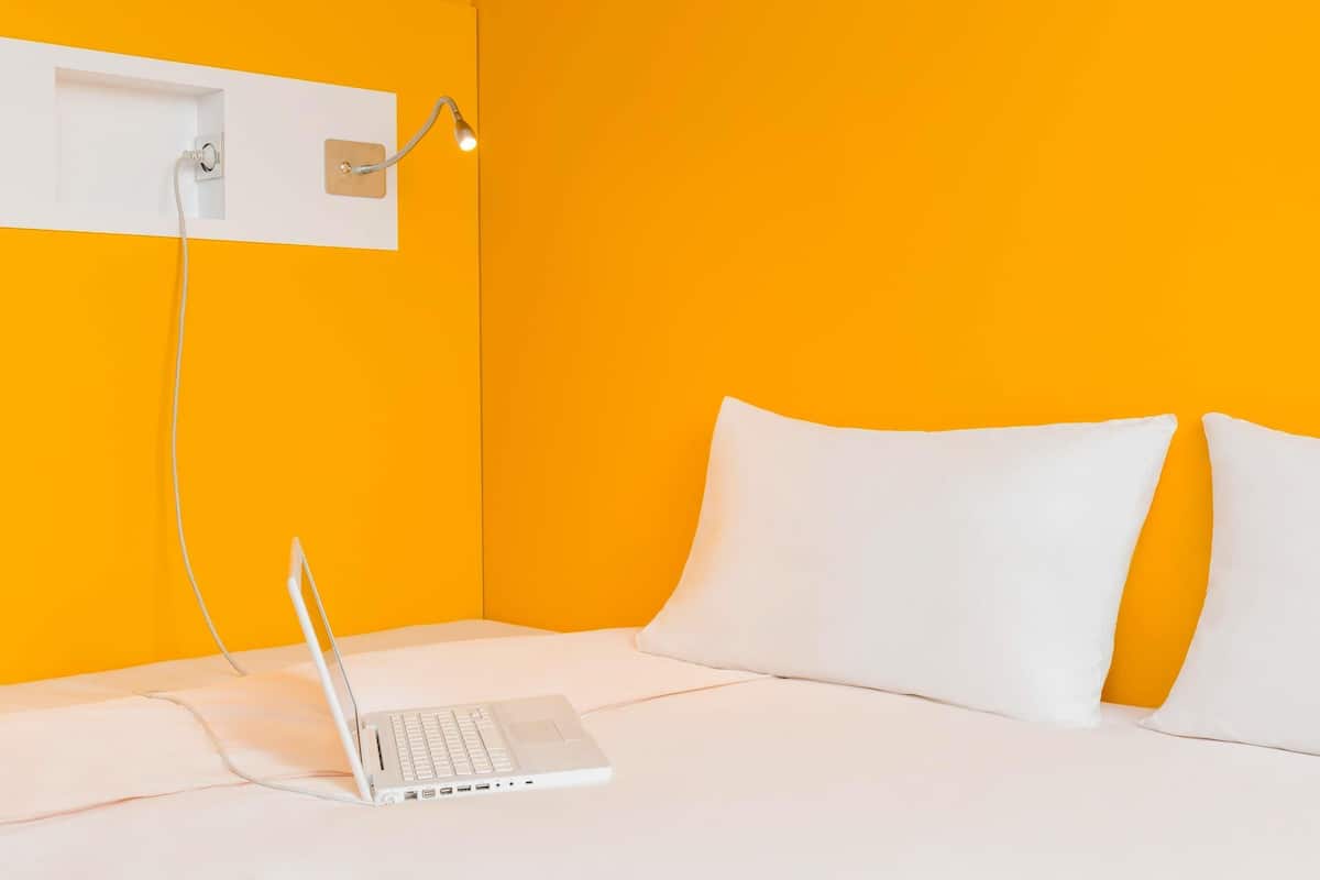 Een kamer met een bed, een laptop op het bed en een wandlamp.