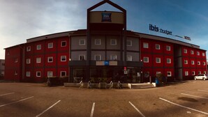 Een meerlaags hotel met een gevel in rood en grijs, een opvallend 'ibis budget' bord en een parkeerplaats ervoor.