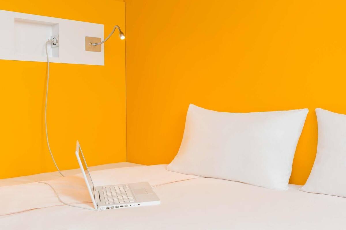 Una habitación con una cama, un ordenador portátil sobre la cama, un enchufe de pared y una lámpara de pared.