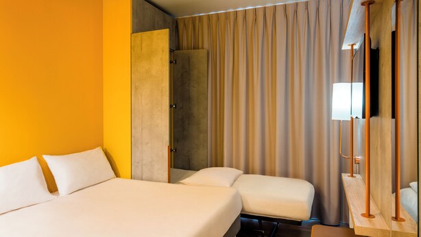 Hotelkamer met een groot bed, een kleiner bed, een houten nachtkastje, een spiegel en een gordijn.
