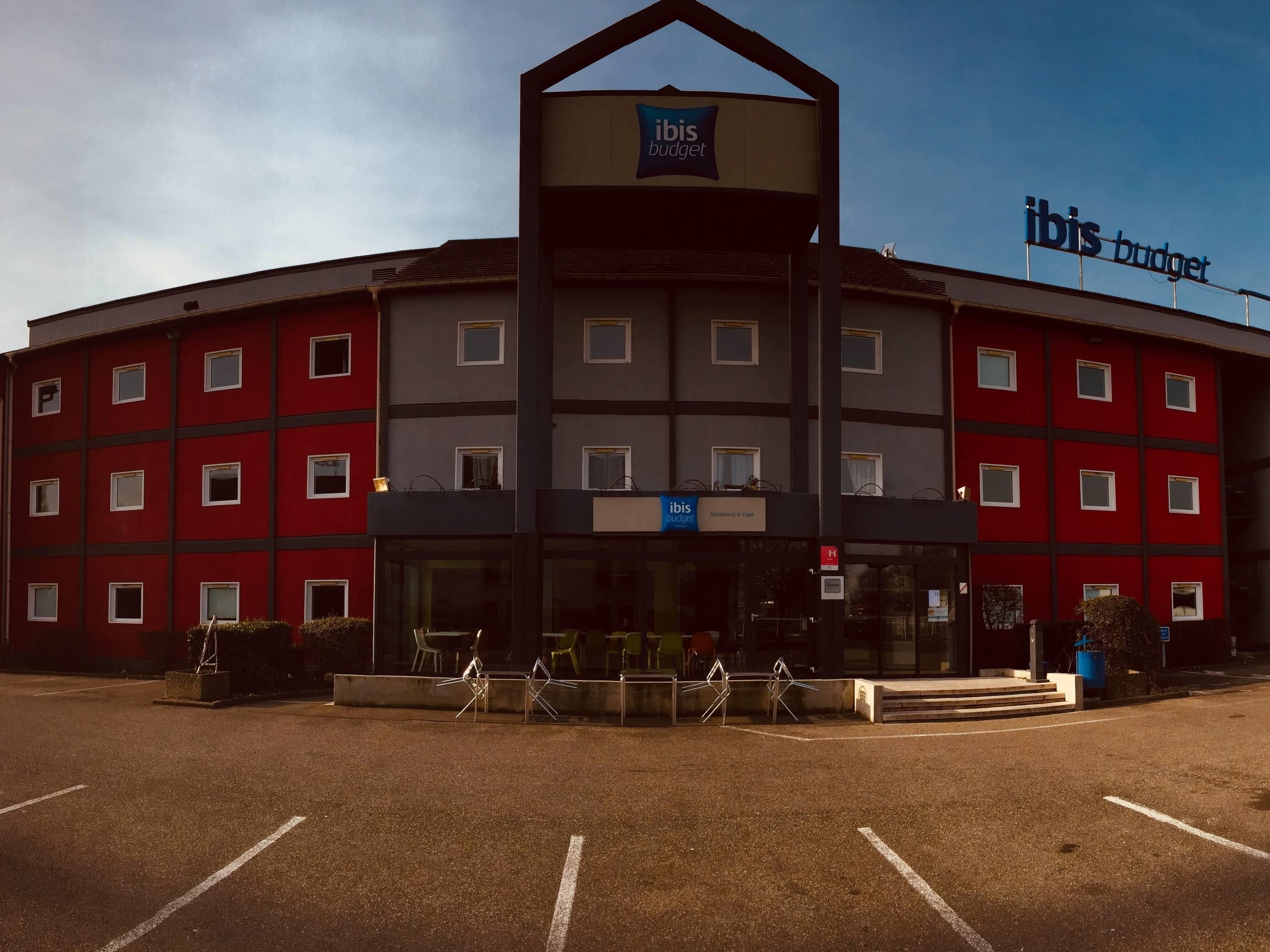 Un hotel de varios pisos con un letrero llamativo de 'ibis budget', un estacionamiento y cielo despejado.