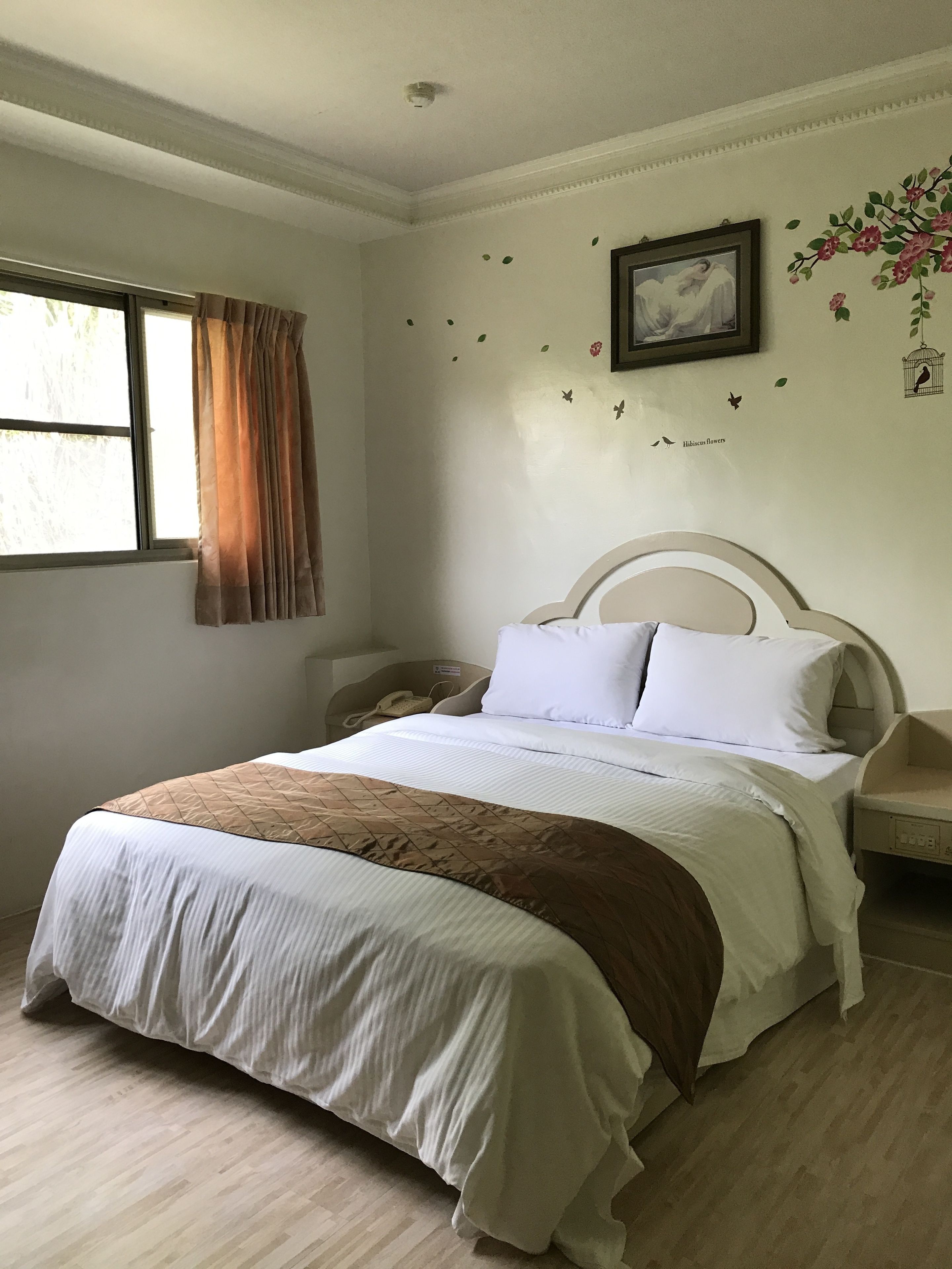 Una cama bien hecha con un edredón marrón, un teléfono junto a la cama y una pared con un cuadro enmarcado y adhesivos decorativos.