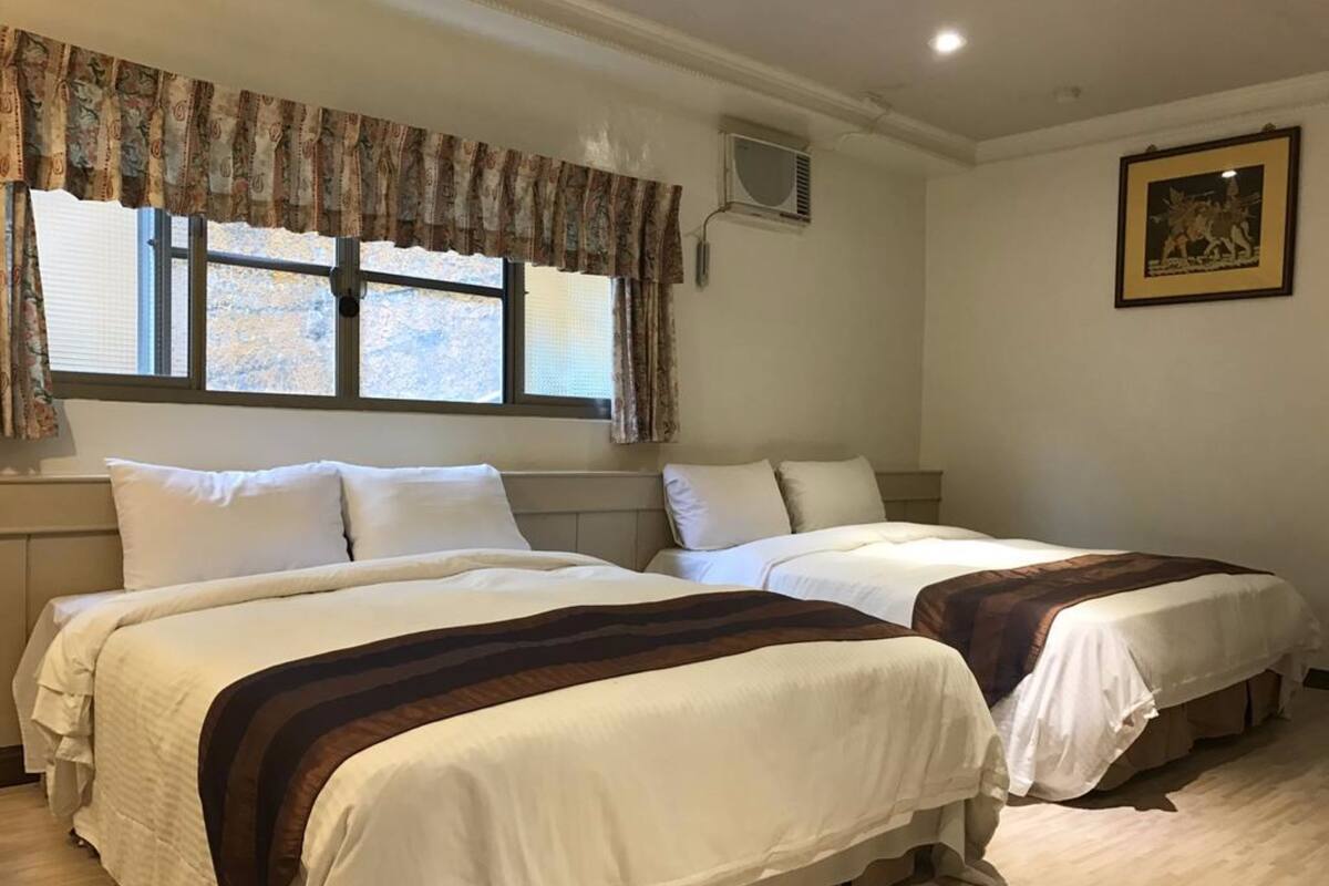 A hotel room with two beds, a window with curtains, an air conditioning unit, and a framed picture on the wall.