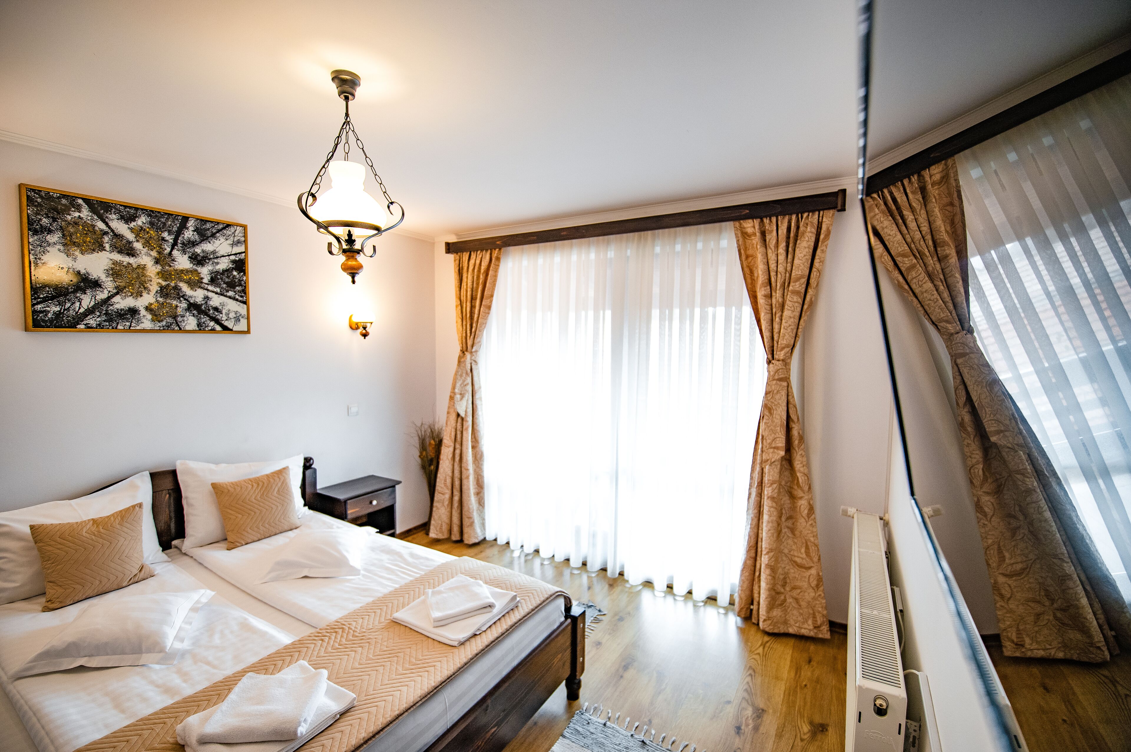 A hotel room with a large bed, a bedside table, a lamp, a radiator, and a window with curtains.