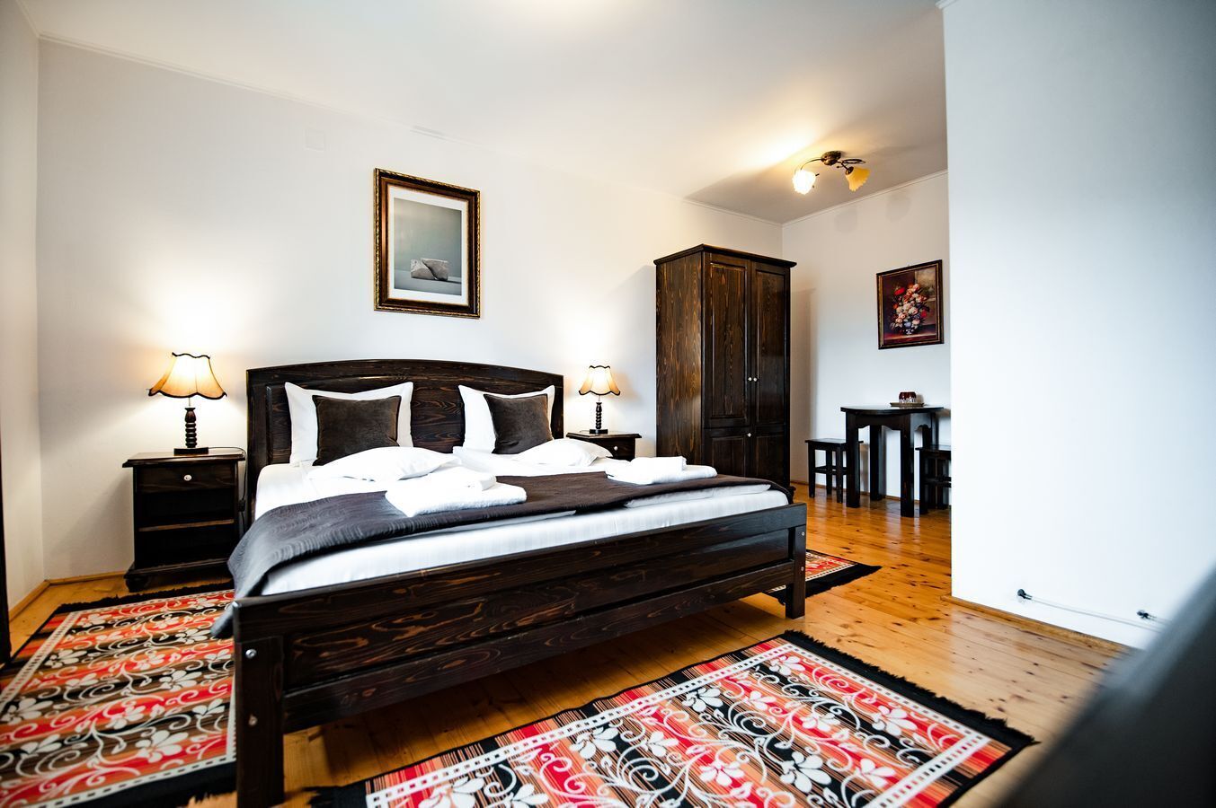 A neatly made bed with a dark wooden frame, two bedside tables with lamps, a wooden wardrobe, and a dining area with a table and chairs.