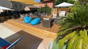 Een terras buiten met houten vloer, zithoeken en een groot geel zonnescherm.