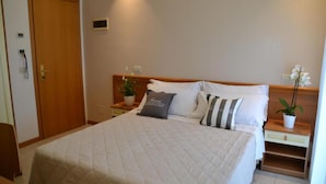A hotel room with a large bed, a wooden headboard, a bedside table with a plant, and a door with a phone. - Apogeo (Rimini)
