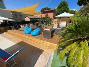 An outdoor patio with wooden flooring, seating areas, and a large yellow shade sail. - Apogeo (Rimini)