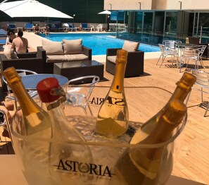 A poolside area with lounge chairs, a glass table, and bottles of Astoria wine in a bucket.