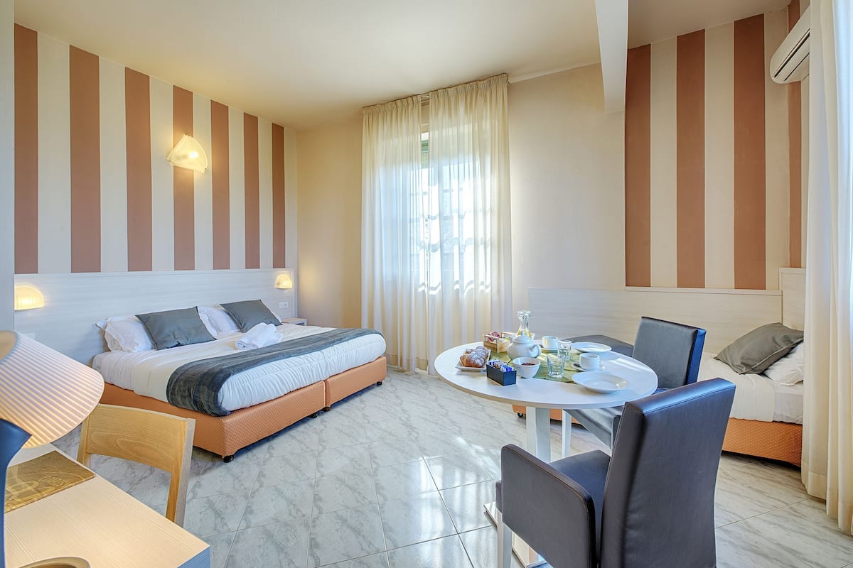 A hotel room with a bed, a dining table with chairs, a sofa, and a window with curtains.