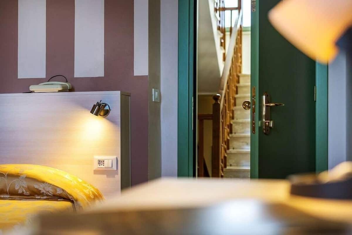 A room with a bed, a bedside table with a lamp, a wall socket, and a door leading to a staircase.