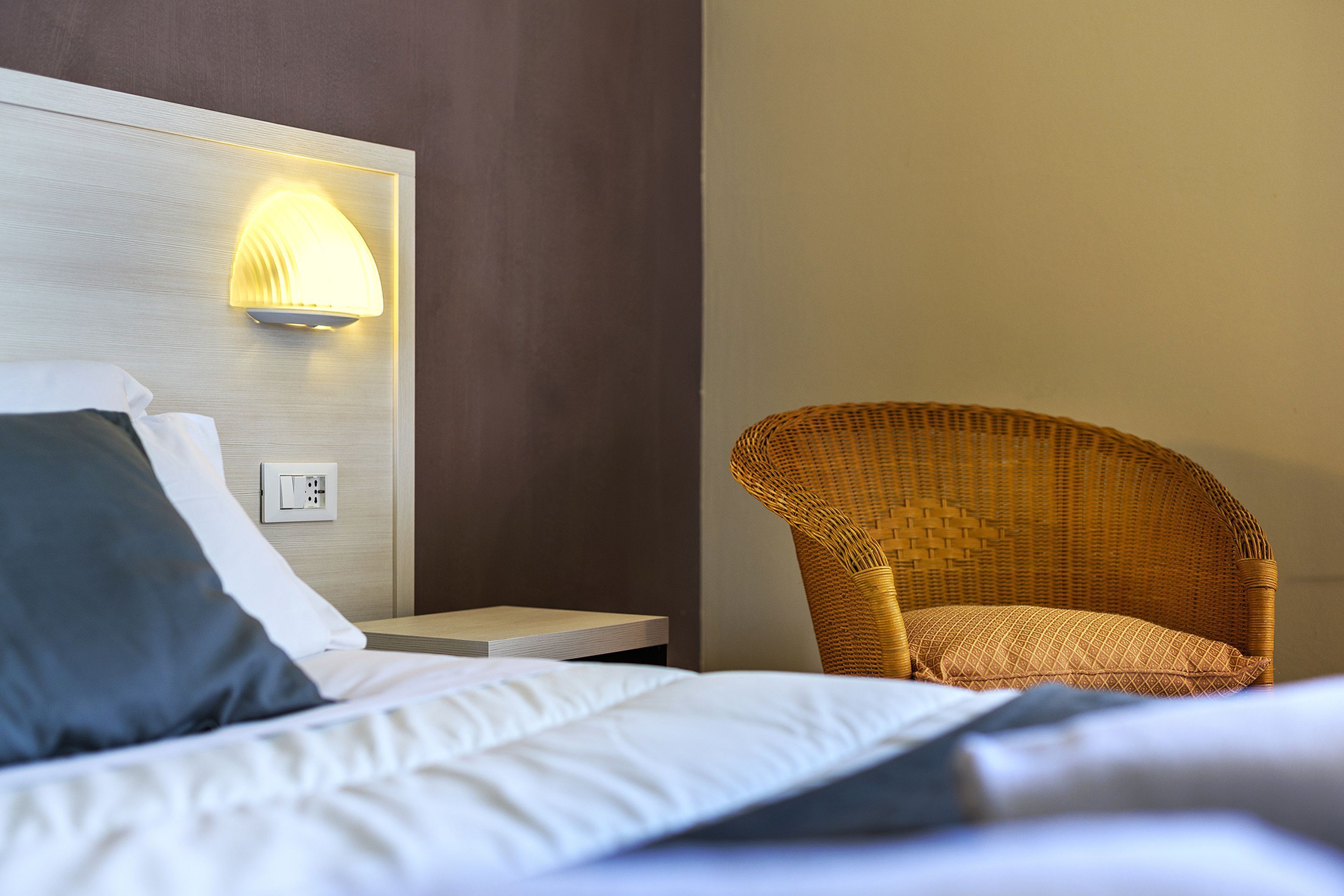 A neatly made bed with pillows, a bedside table, a wall-mounted light, and a wicker chair.