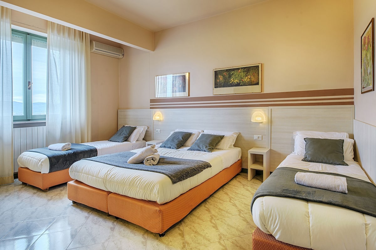 A hotel room with three beds, a window with curtains, an air conditioner, and two wall-mounted lights.