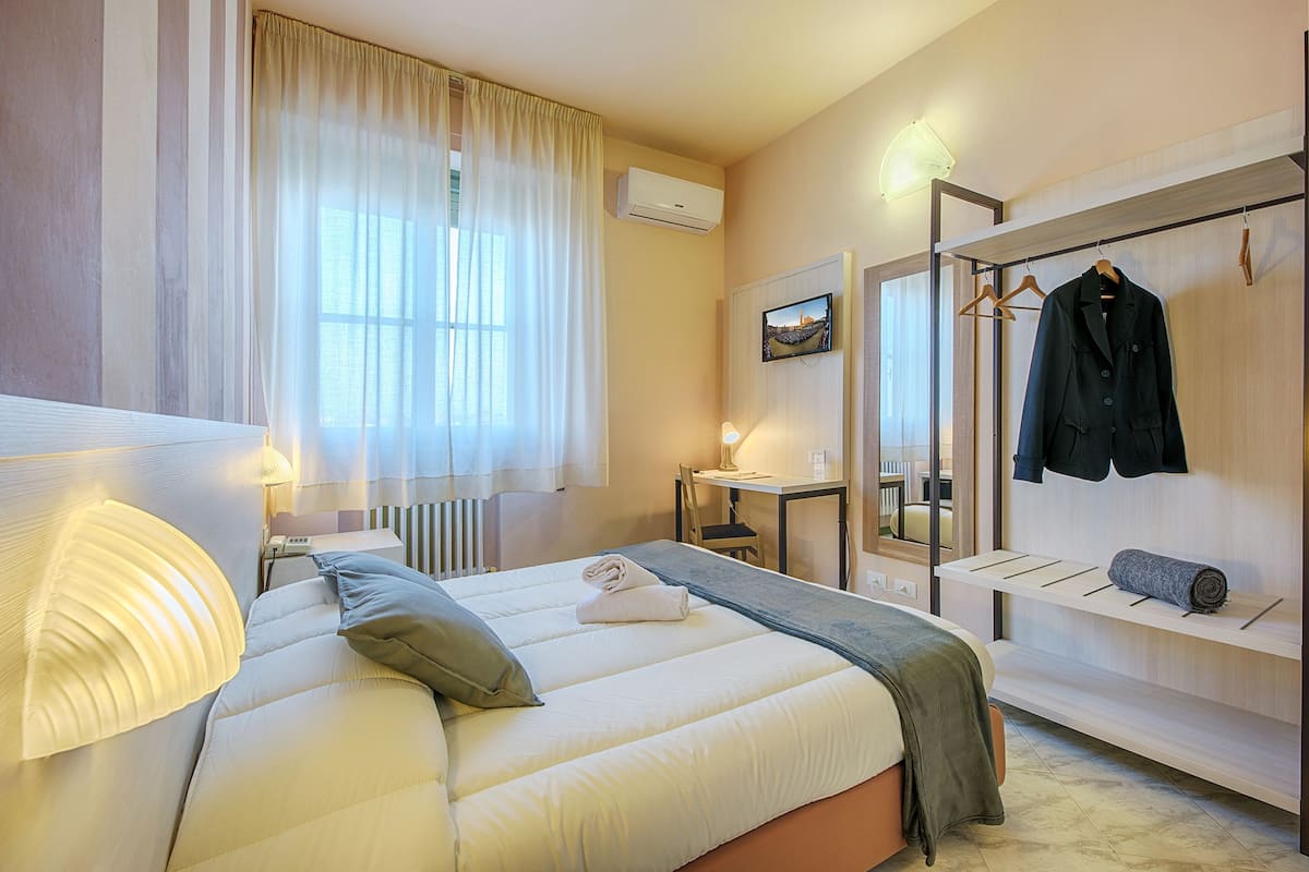 A hotel room with a bed, a desk, a chair, a mirror, and a coat rack.