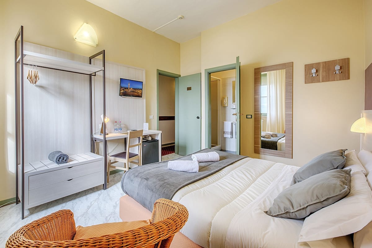 A hotel room with a bed, a desk, a chair, a wardrobe, and a bathroom visible through an open door.