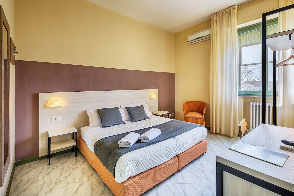 A hotel room with a large bed, bedside tables, a chair, a desk, and a window with curtains.