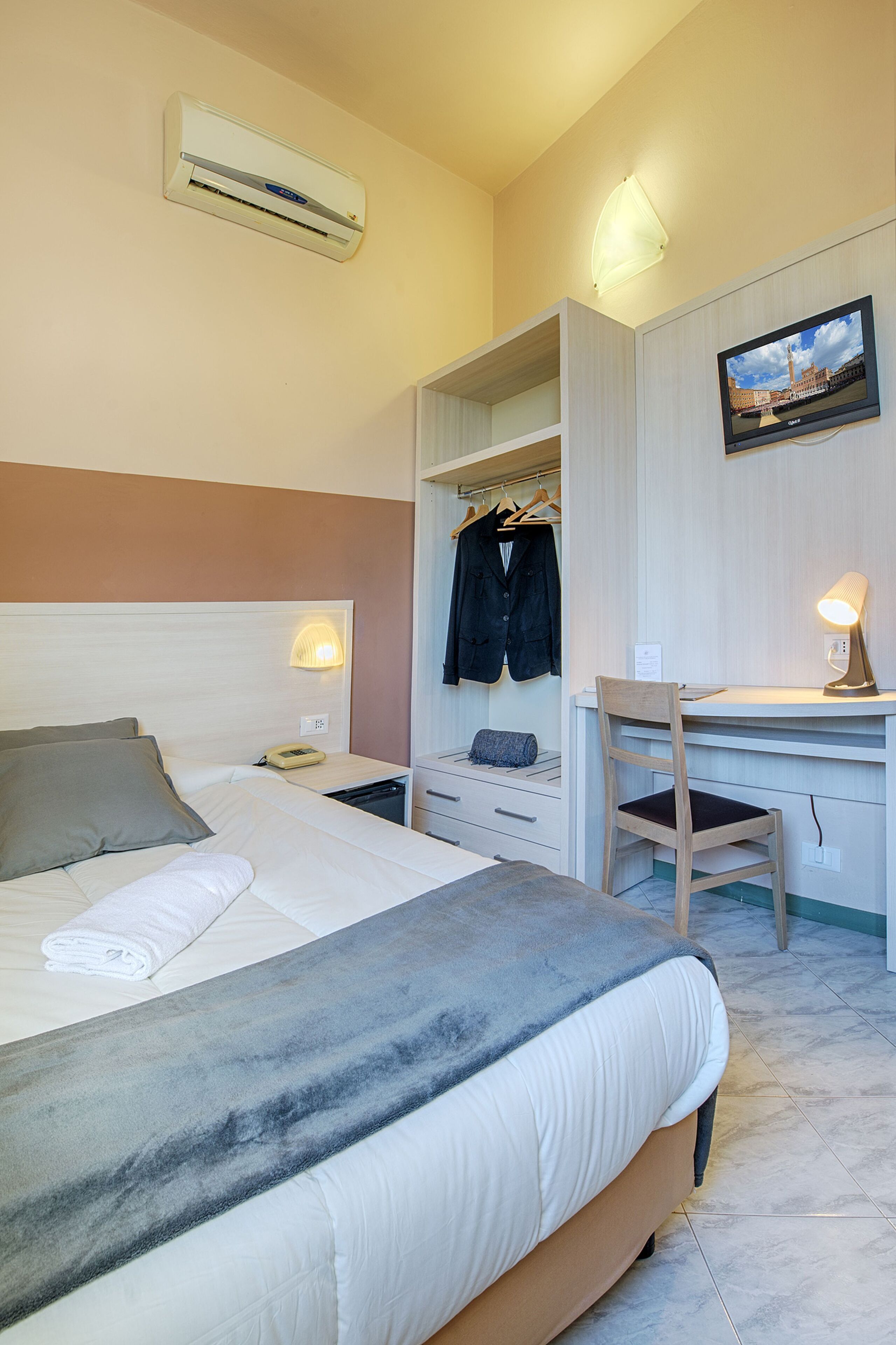 Ein Hotelzimmer mit Bett, Nachttisch, Lampe, Schreibtisch mit Stuhl, Kleiderschrank und Fernseher an der Wand.