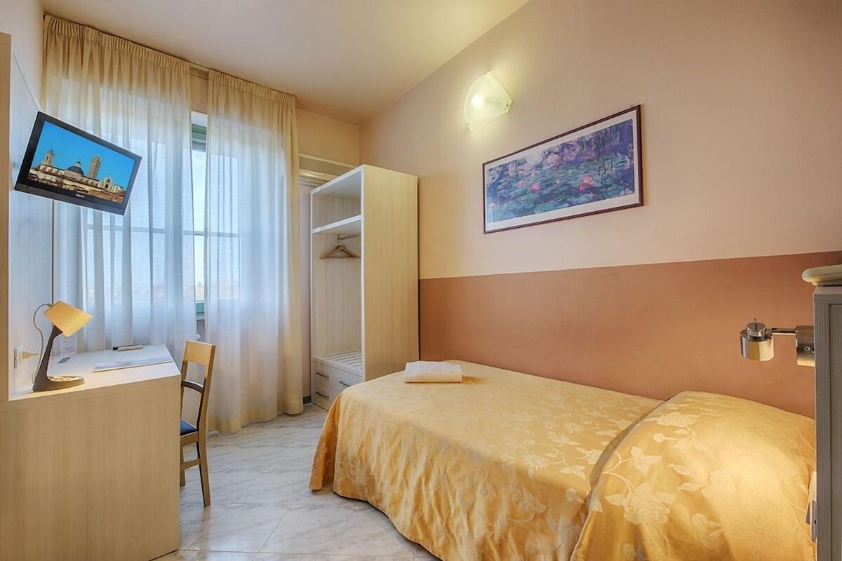 A hotel room with a bed, a desk with a lamp, a TV, a painting, and a wardrobe.