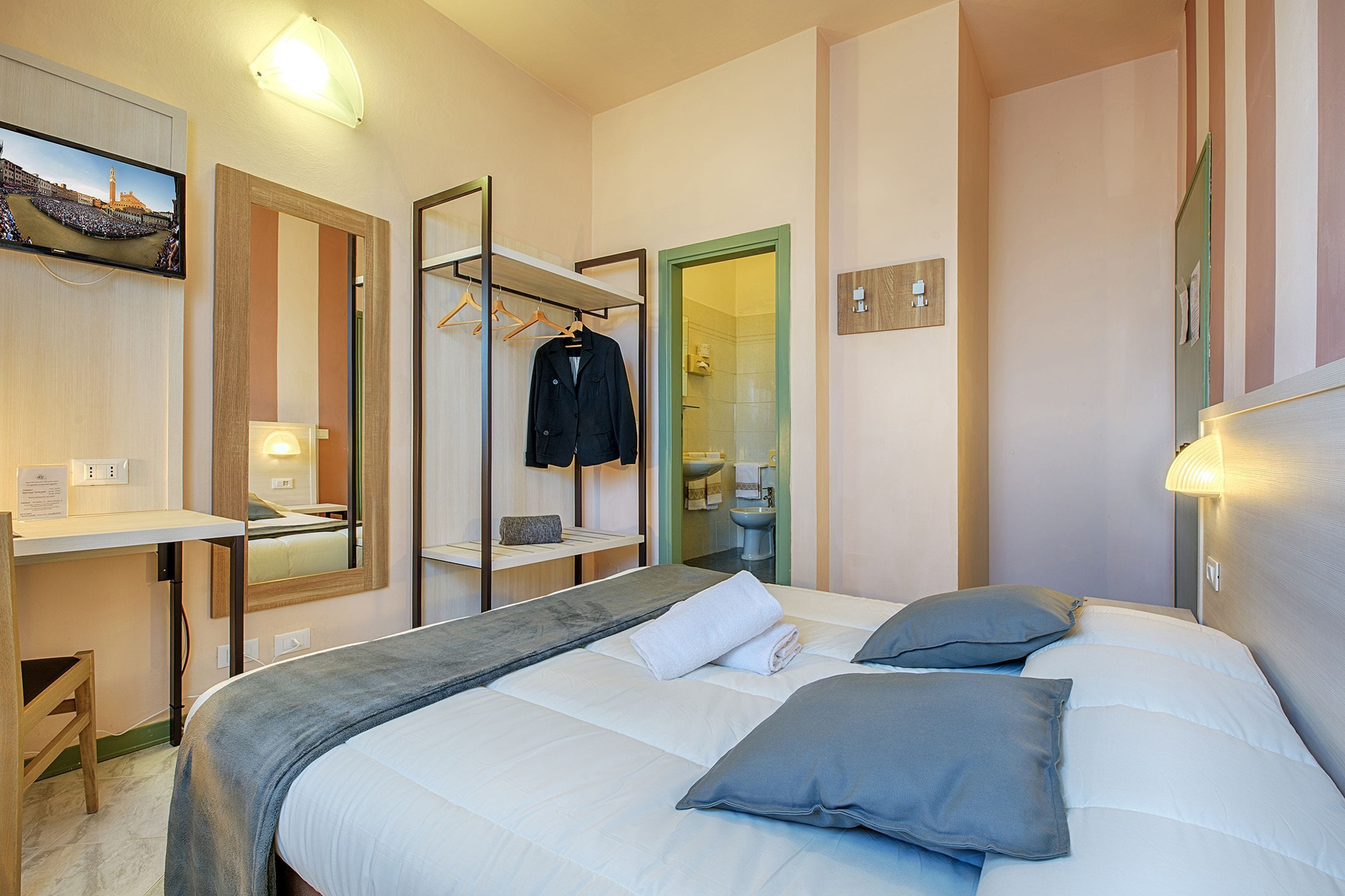 A hotel room with a bed, a desk, a mirror, a TV, and a bathroom visible through an open door.