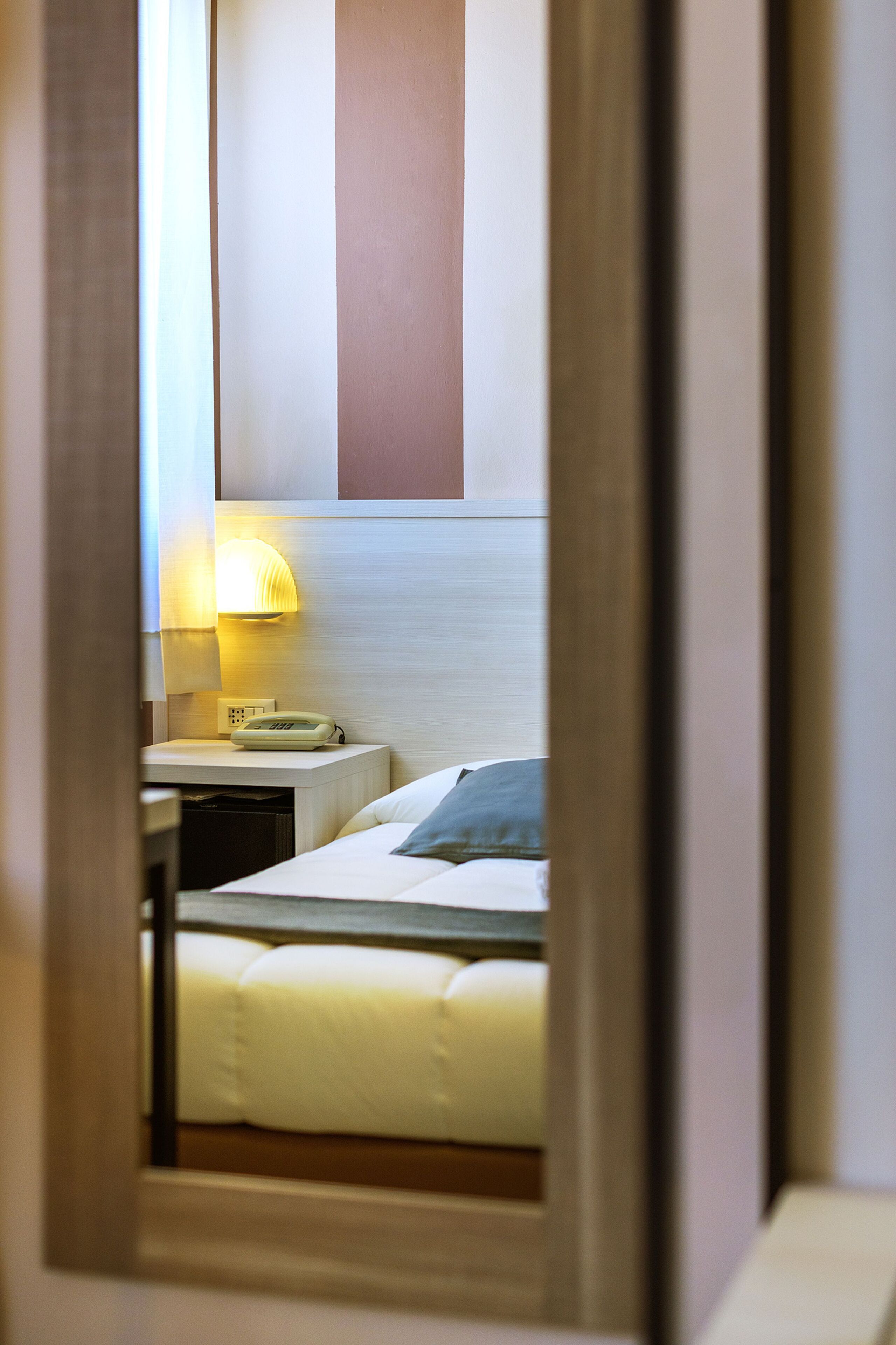 A hotel room with a bed, a bedside table with a lamp, and a mirror reflecting the room.