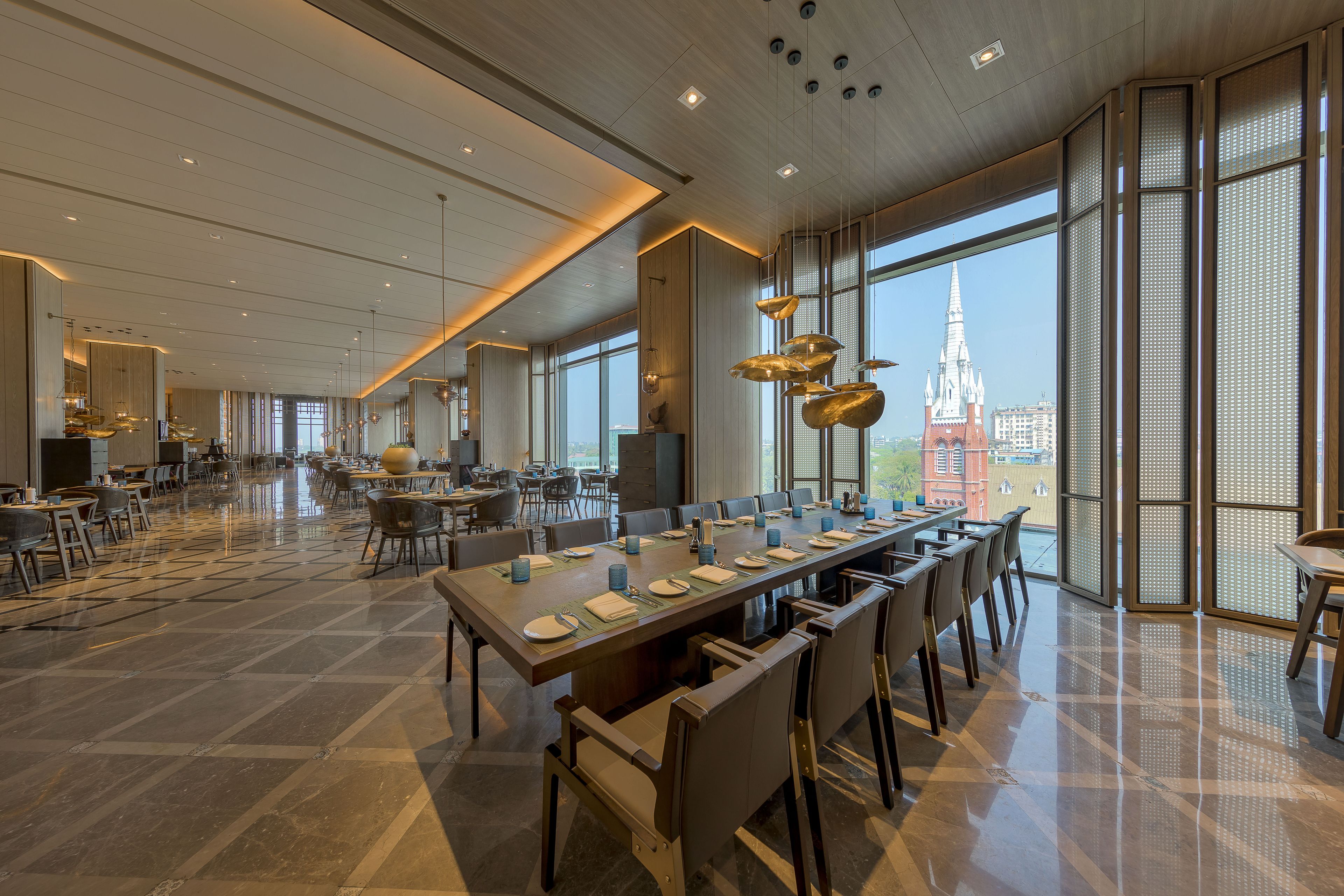 A spacious dining area with a long table set for a meal, large windows offering a view of a cityscape, and modern lighting fixtures.