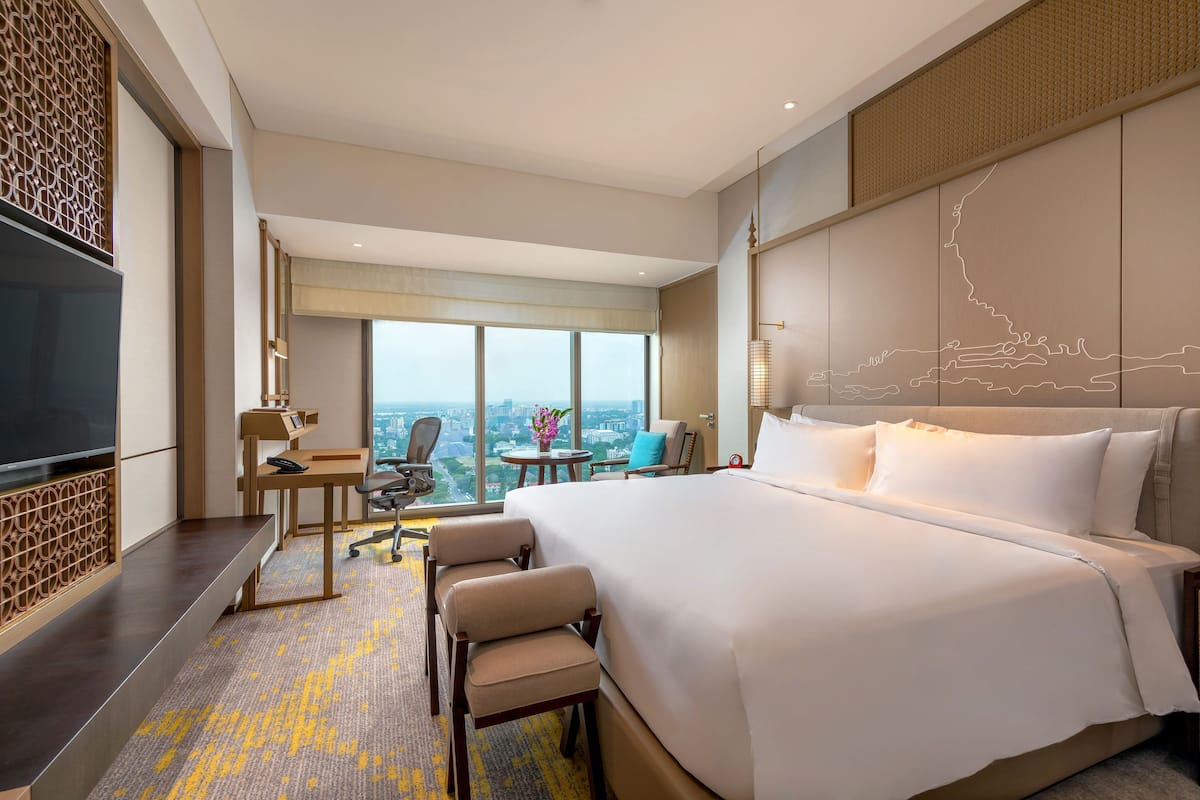 A modern hotel room with a large bed, a desk with a chair, a television, and a view of the city.