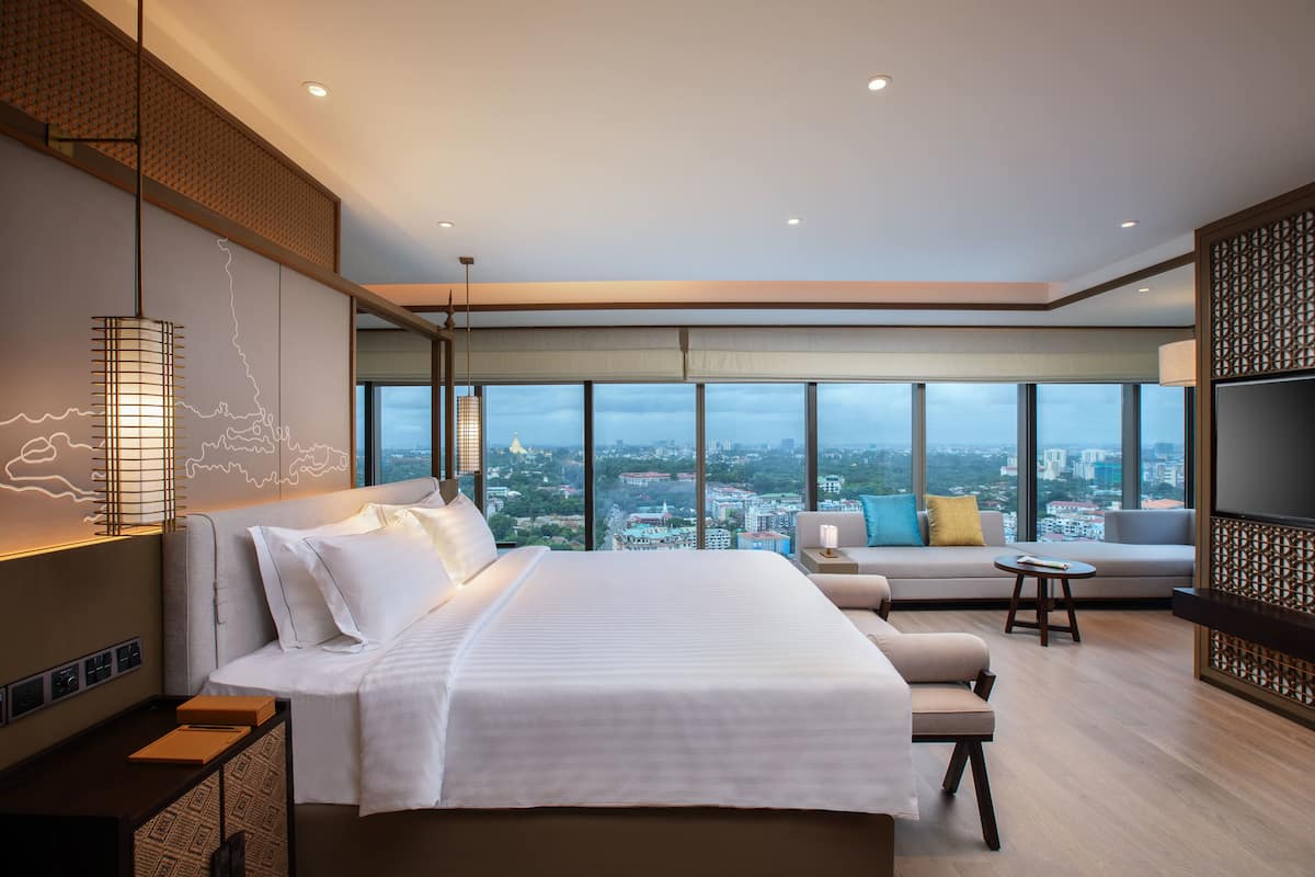 A modern hotel room with a large bed, a sofa, a TV, and a city view.