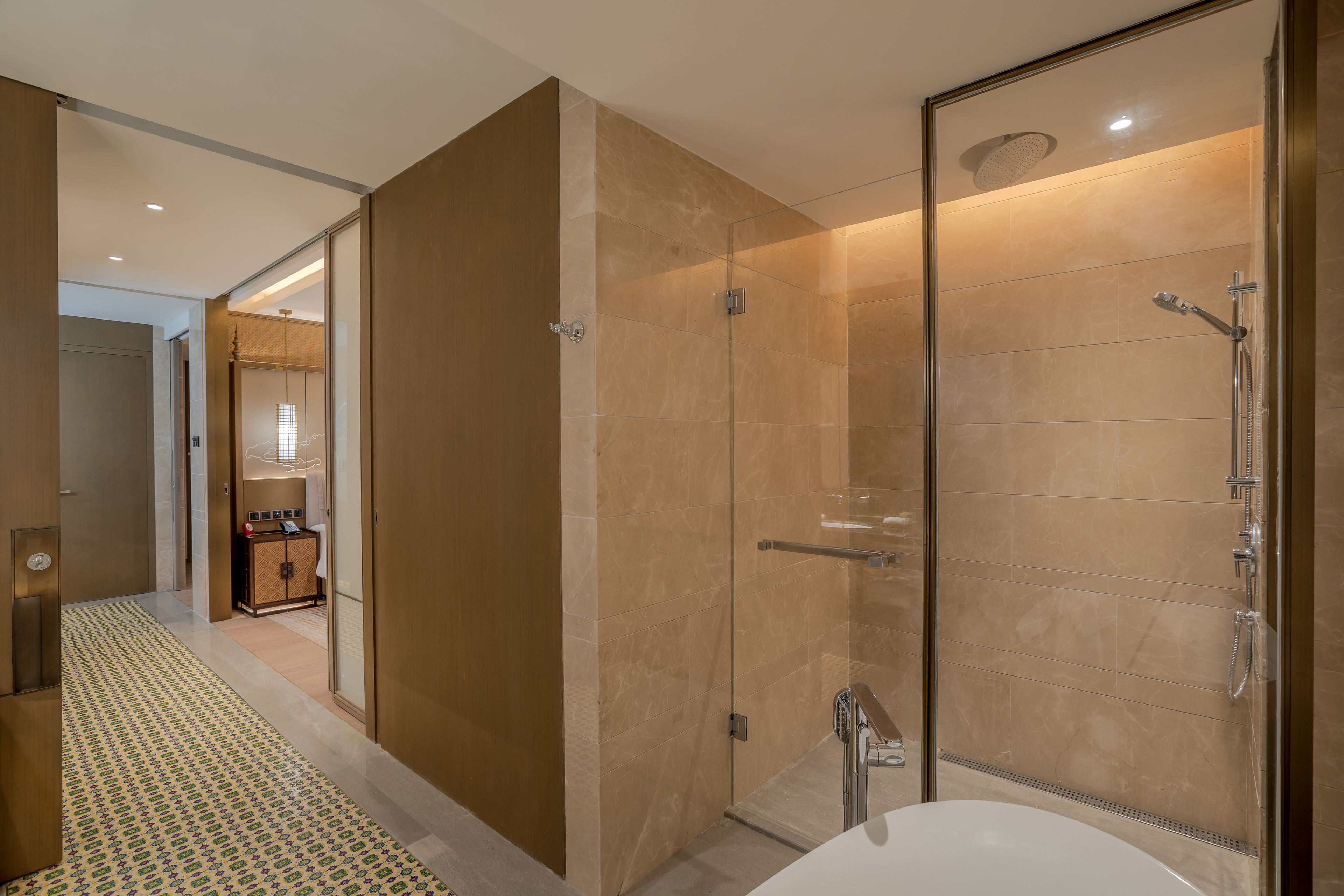 A modern bathroom with a glass-enclosed shower, a large bathtub, and a patterned hallway leading to a room with a minibar.