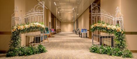 A hotel corridor with a geometric patterned carpet, white metal gates with floral decorations, and tables with artwork on either side.