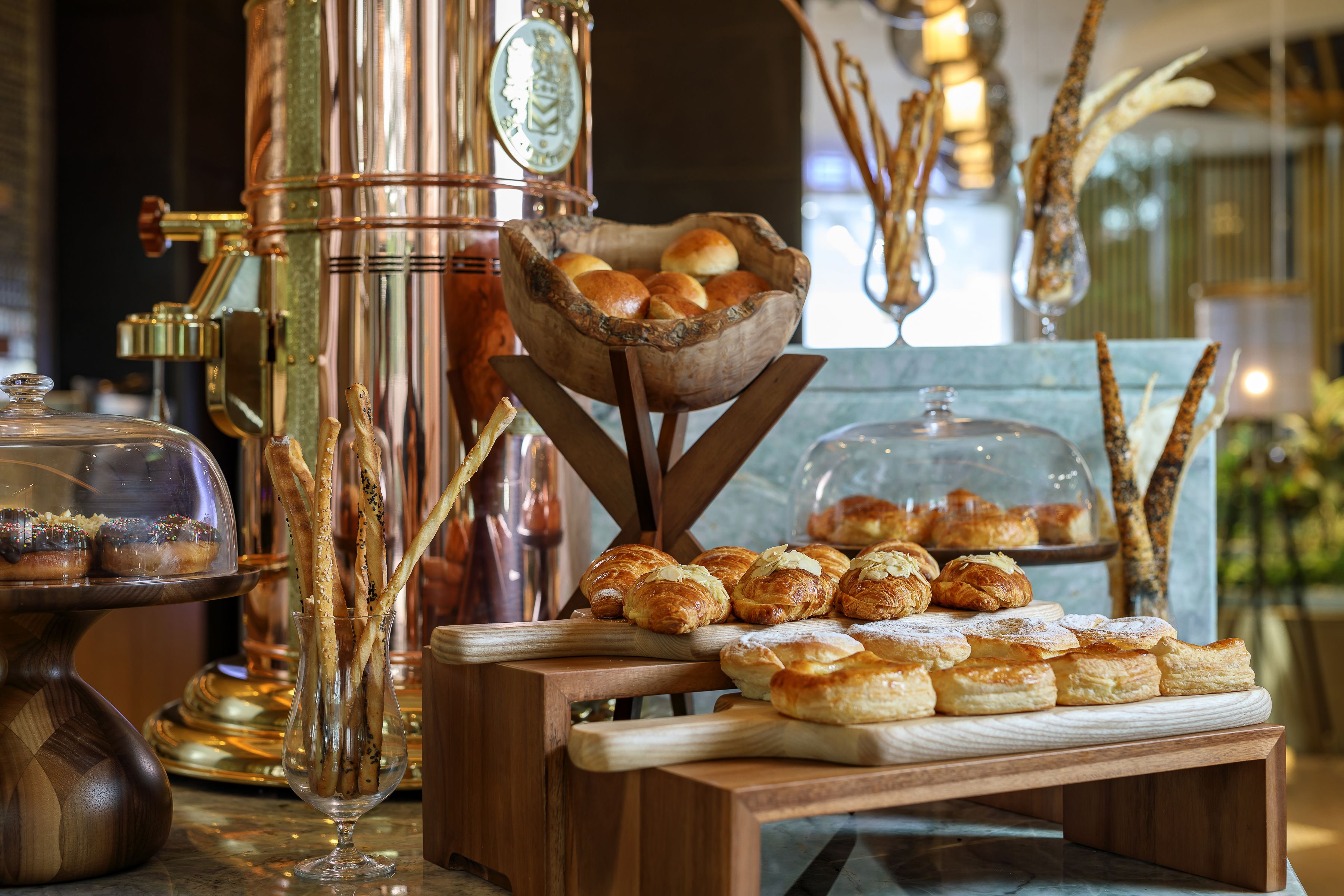 Mesa de buffet com pães, bolos e uma máquina de café, além de uma tigela de madeira decorativa.