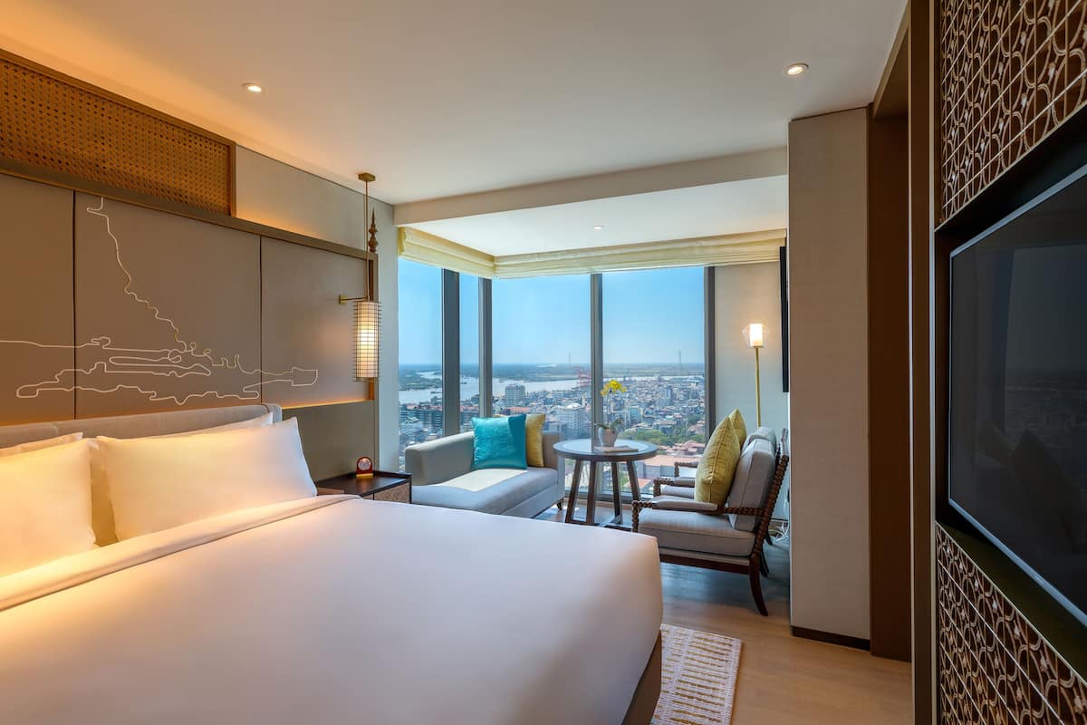 A hotel room with a large bed, a sofa, a chair, a TV, and a view of the city.